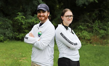 Two smiling people, wearing white long-sleeve shirt, stand back to back with their arms crossed.